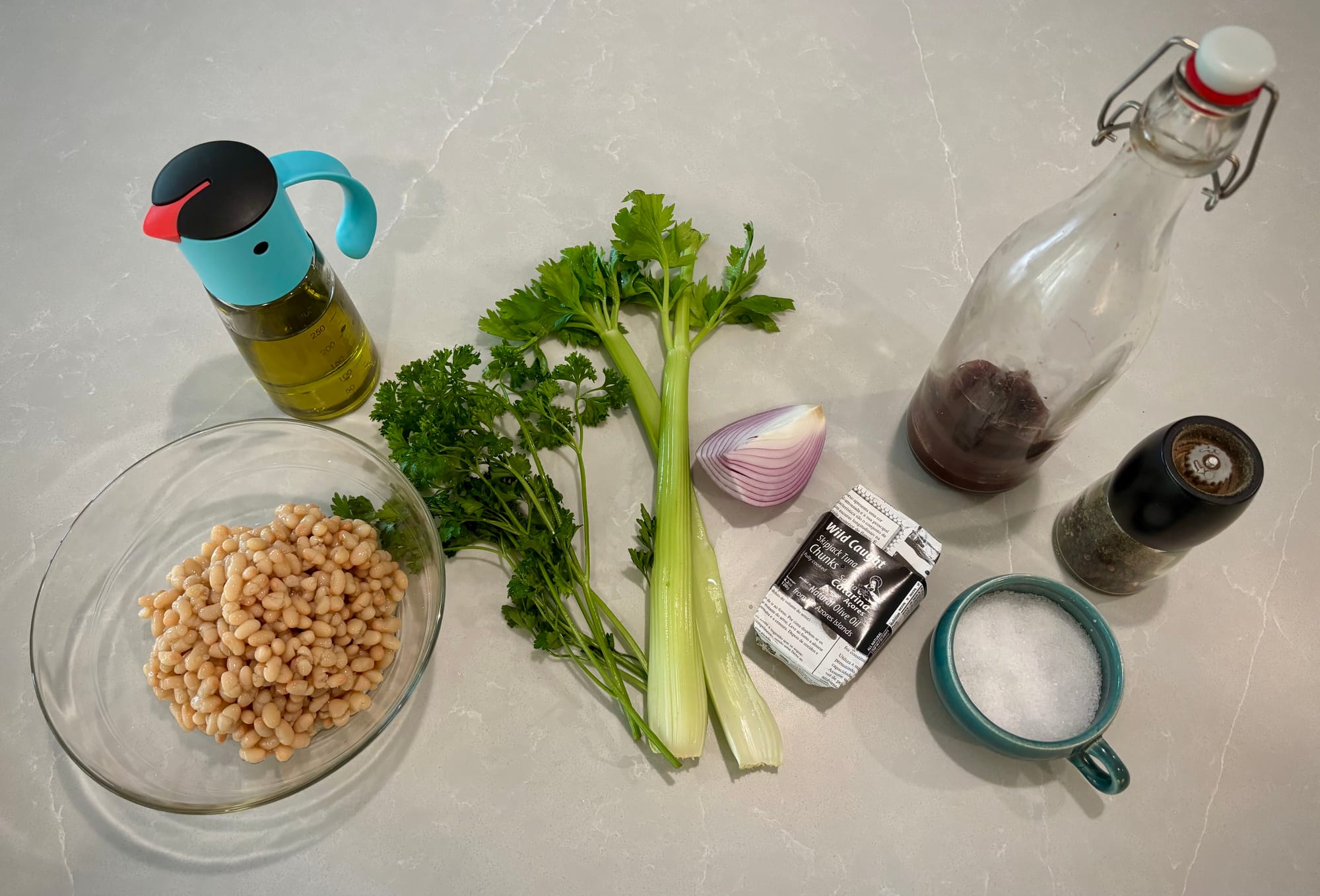 Ingredients for bean salad: beans, olive oil, parsley, celery, red onion, canned tuna, vinegar, pepper and salt, laid out on a countertop