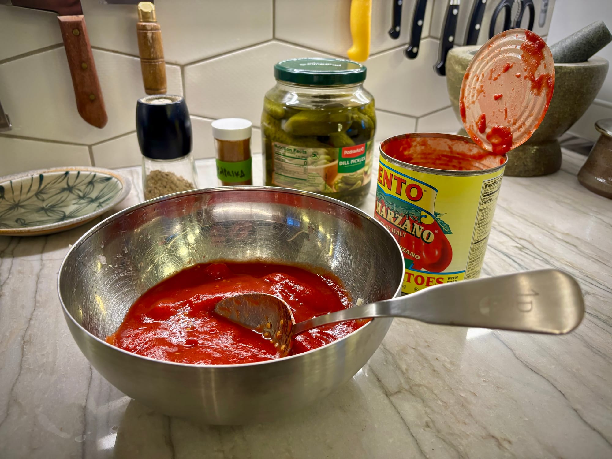 A bowl of canned tomatoes, plus containers of pickles, paprika and white pepper.